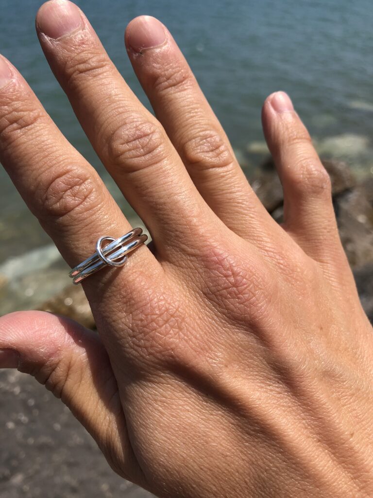 a sterling silver ring of stacked bands joined by a jump ring, worn on a hand held up in front of a body of water 