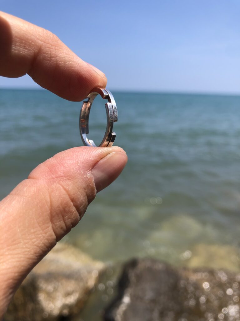 squared silver ring held between two fingers against a background of blue sky and lake